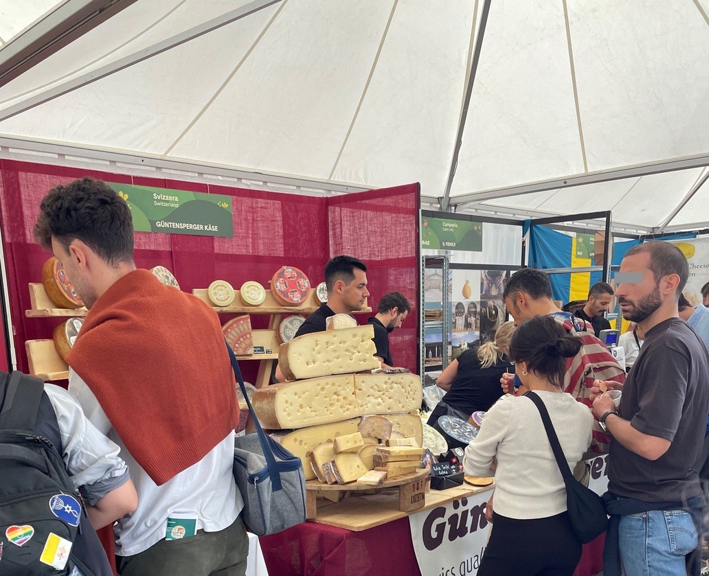 Uno stand di formaggi svizzeri con grossi formaggi e un po’ di gente davanti.