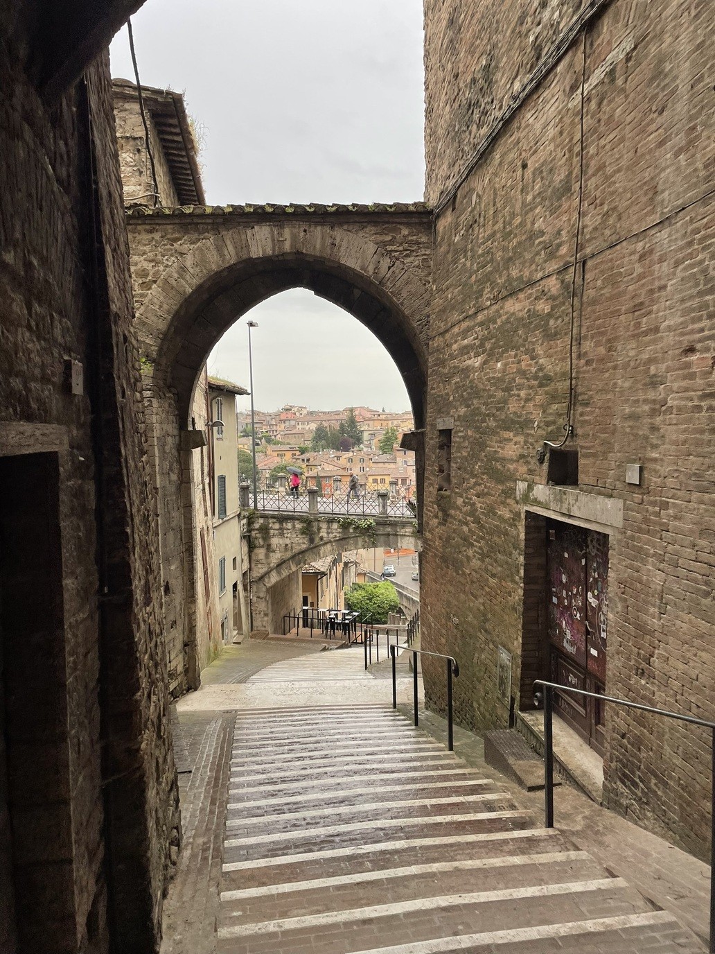 Scale tra vecchi palazzi che passano sotto un ponte, con un cielo grigio.